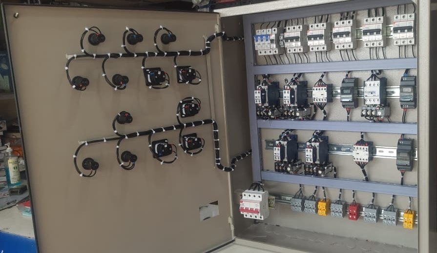The inside of a control panel: rows of MCBs on DIN rail, contactors and overload relays, terminal blocks along the base, and spiral-wrapped wiring looms running to the door-mounted controls